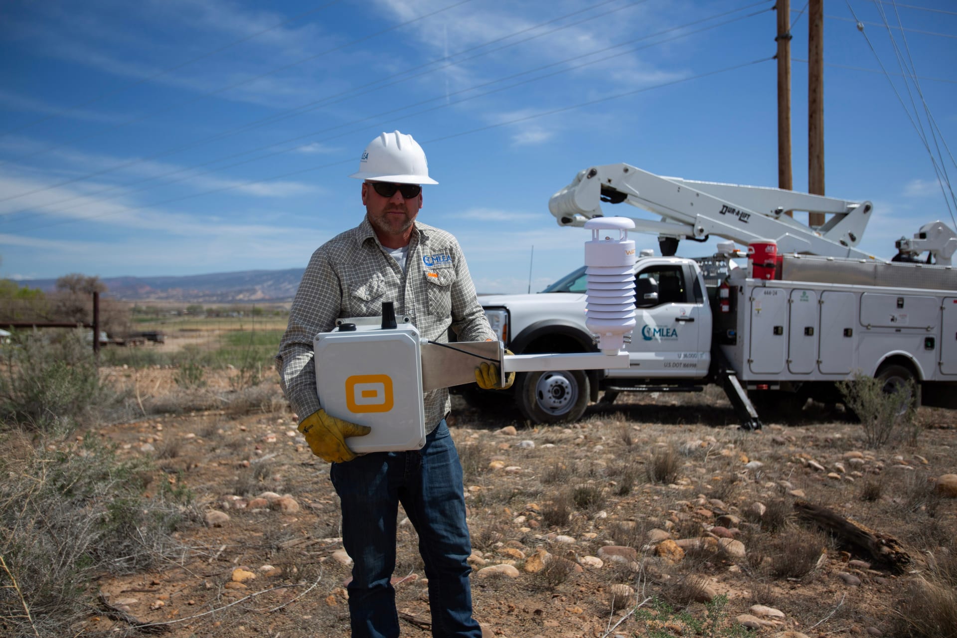 WatchPost technician in a hard hat holding a sensor unit beside a utility service truck, open landscape behind
