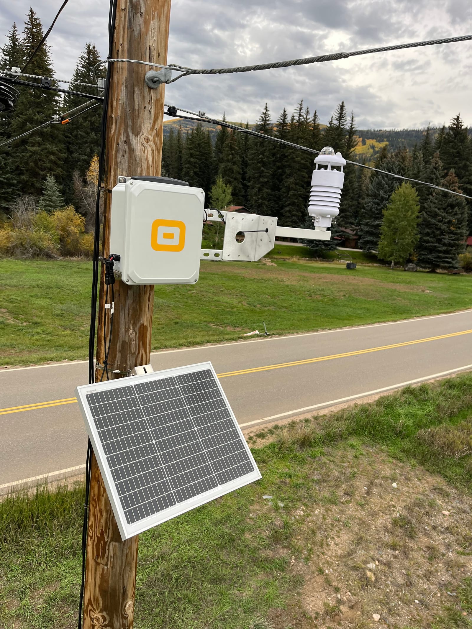 WatchPost weather station with its sensor arm and solar panel mounted on a utility pole beside a rural mountain road