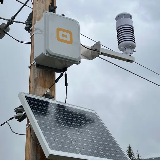 A WatchPost weather station deployed on a utility pole, with its solar panel and sensor mast against a mountain sky